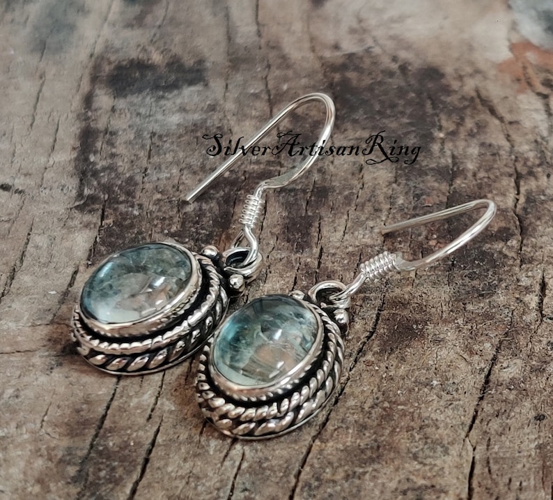 May include: Silver earrings with a blue gemstone set in a silver bezel with a rope-like design. The earrings have a hook closure.