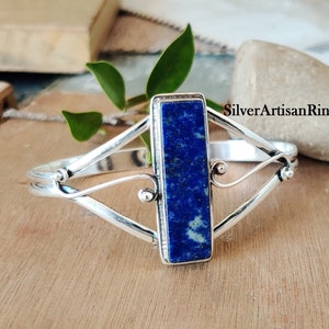 Könnte beinhalten: Ein silbernes Armband mit einem blauen Lapislazuli-Stein in der Mitte. Das Armband hat ein filigranes, kompliziertes Design mit einem gedrehten Drahtmuster. Der Stein ist rechteckig und hat eine glatte, polierte Oberfläche. Das Armband ist ein wunderschönes und einzigartiges Schmuckstück.