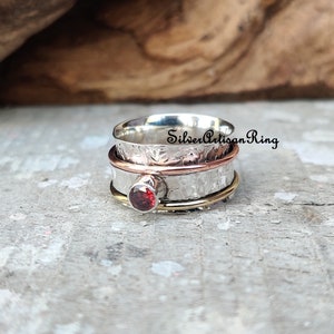 May include: A silver, copper, and gold spinning ring with a red gemstone. The ring is textured and has a hammered finish. "SilverArtisanRing" is written on the ring.