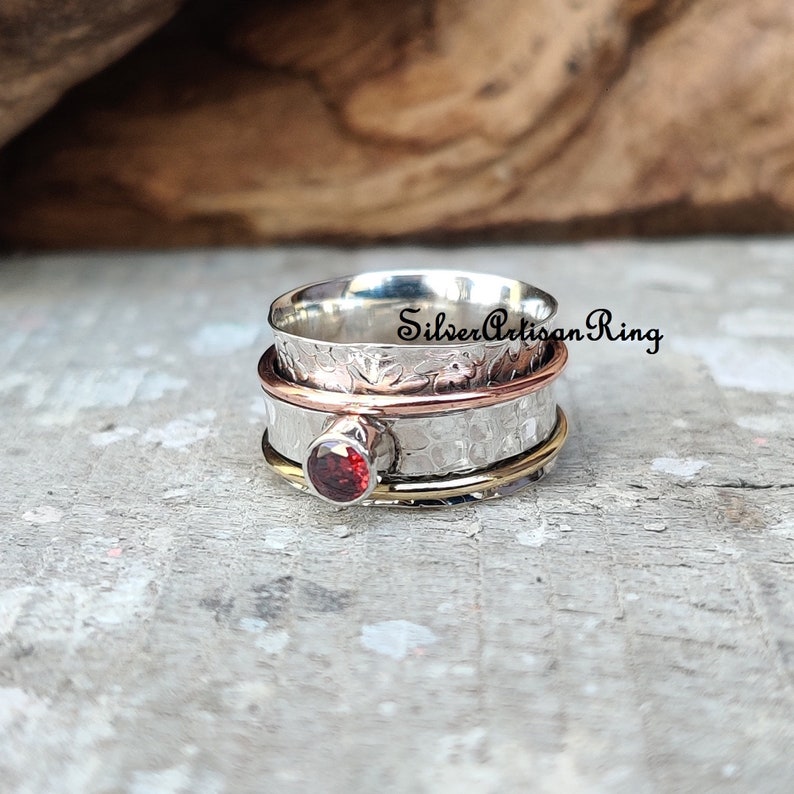 May include: A silver, copper, and gold spinning ring with a red gemstone. The ring is textured and has a hammered finish. "SilverArtisanRing" is written on the ring.