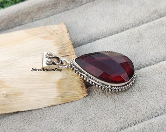 Puede incluir: Un colgante de plata con una piedra preciosa de color rojo oscuro en forma de lágrima. El colgante tiene un borde de plata detallado y un aro para sujetar una cadena.