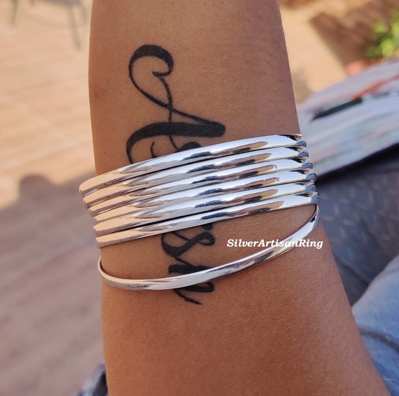 May include: A collection of polished silver bangles. The bracelets have a smooth, reflective surface and a minimalist design. The bangles are stacked, creating a layered effect. The jewellery is a timeless accessory, suitable for various occasions.