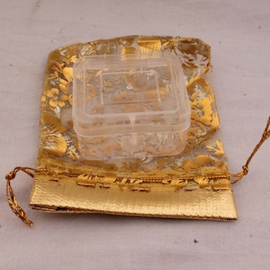 May include: A clear plastic square box with a lid, inside a gold and white floral patterned drawstring bag.