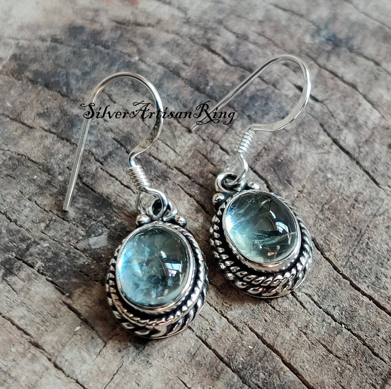 May include: A pair of silver earrings with blue aquamarine gemstones set in a detailed silver bezel. The earrings have a simple hook closure.