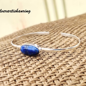 Könnte beinhalten: Ein silbernes Armreif-Armband mit einem blauen, ovalen Lapislazuli-Edelstein. Das Armband liegt auf einer braunen gewebten Oberfläche.