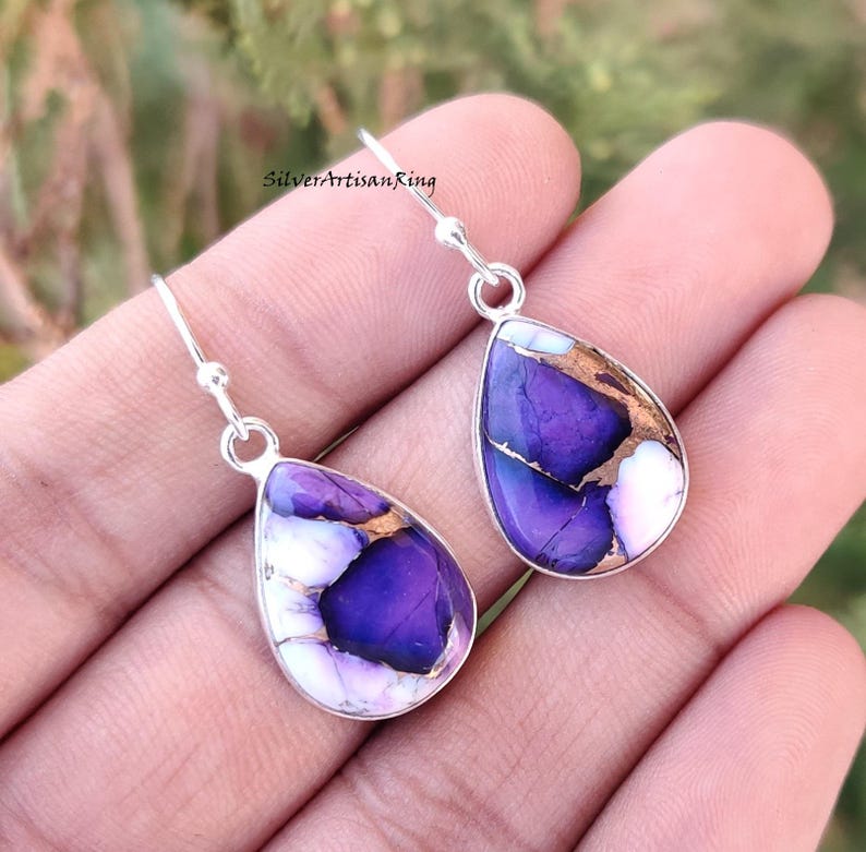 Pu&ograve; includere: Un paio di orecchini pendenti in argento con gocce di pietre preziose viola e bianche. Le pietre preziose hanno un motivo marmoreo con accenti dorati.