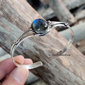 Puede incluir: Un brazalete de plata con una piedra preciosa de labradorita azul. El brazalete tiene un dise&ntilde;o delicado con dos bandas curvas y peque&ntilde;as cuentas.
