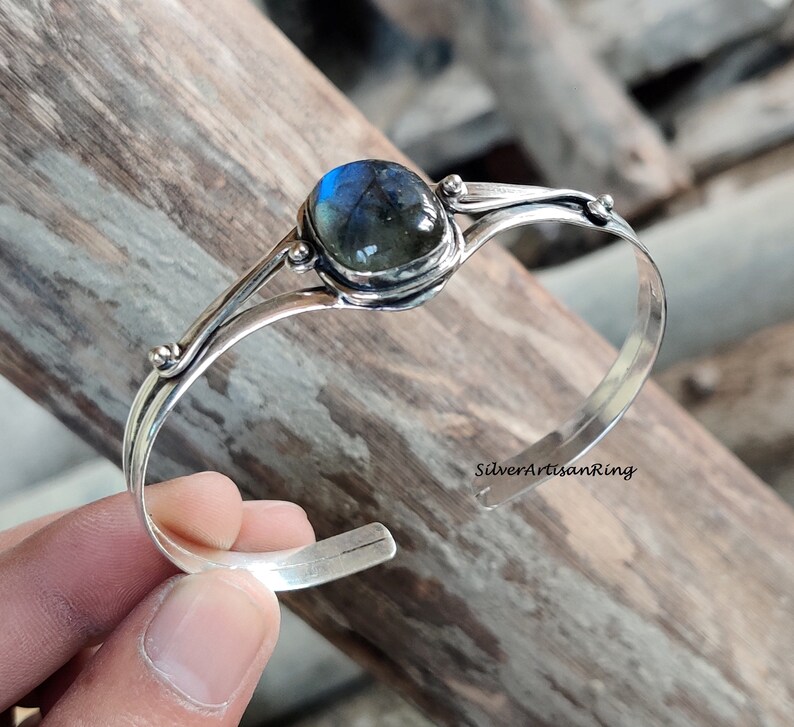 Puede incluir: Un brazalete de plata con una piedra preciosa de labradorita azul. El brazalete tiene un dise&ntilde;o delicado con dos bandas curvas y peque&ntilde;as cuentas.