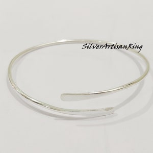 Op de afbeelding: Een zilverkleurige, open manchetarmband met een minimalistisch ontwerp. De armband heeft een gladde, gepolijste afwerking en heeft een open ontwerp met twee platte, afgeronde uiteinden. De tekst "SilverArtisanRing" is zichtbaar.