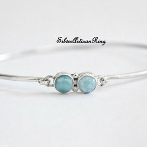 May include: A silver bangle bracelet with two light blue gemstone accents. The bracelet is open and has a clasp. The text "Silver Artisan Ring" is visible in the image.