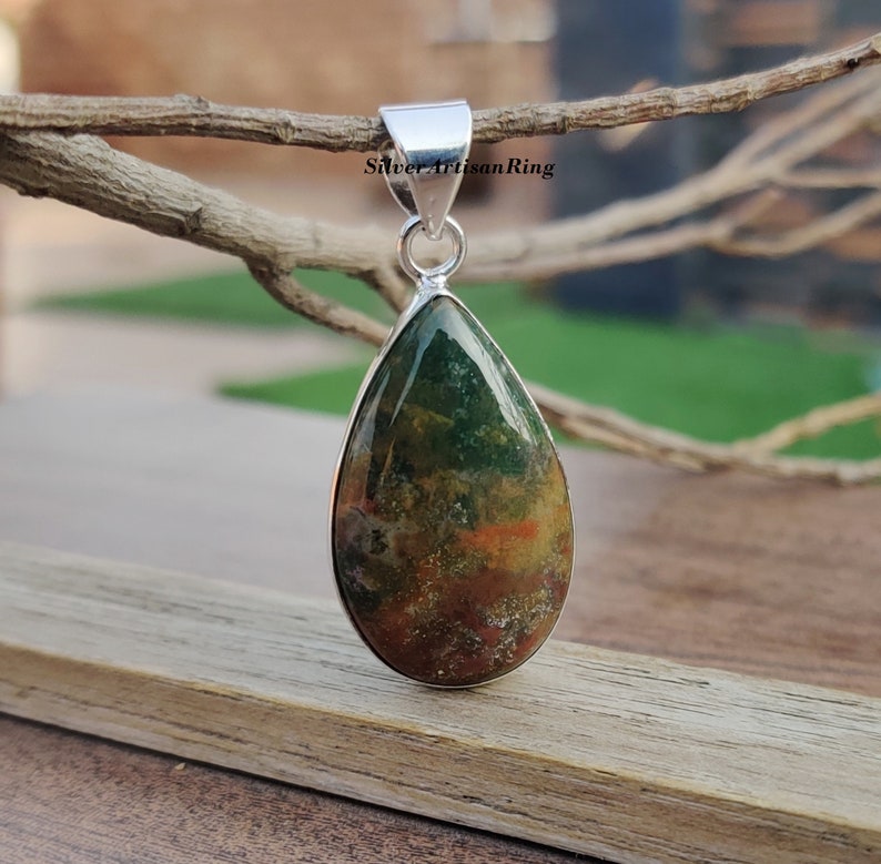 May include: A teardrop-shaped pendant with a green, brown, and orange moss agate stone set in sterling silver. The pendant is hanging from a branch.