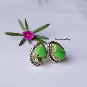 May include: A pair of teardrop-shaped stud earrings with a bright green and brown stone set in a silver-colored metal. The earrings have a textured border. A small pink flower and green leaves are in the background.