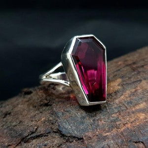 Könnte beinhalten: Ein silberner Ring mit einem großen, facettierten, dunkelrosa Edelstein in Form eines Sarges.