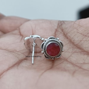 Könnte beinhalten: Silberne Ohrstecker mit einem roten Edelstein in der Mitte. Die Ohrringe haben ein detailliertes, verziertes Design um den Edelstein herum. Der Stift ist sichtbar, vor neutralem Hintergrund.