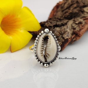 Könnte beinhalten: Ein silberner Ring mit einer weißen Kaurischnecke in der Mitte. Der Ring ist von einem Perlenrand umgeben.