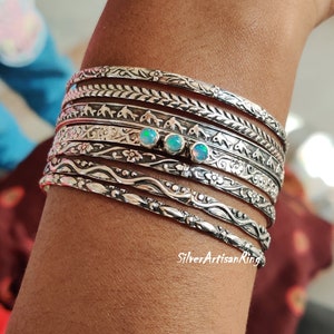 Könnte beinhalten: Ein Set aus acht silbernen Armreifen mit aufwendigen Designs, darunter florale Muster, seilartige Texturen und geometrische Formen. Drei kleine Opal-Steine ​​sind in der Mitte der Armreifen eingefasst.