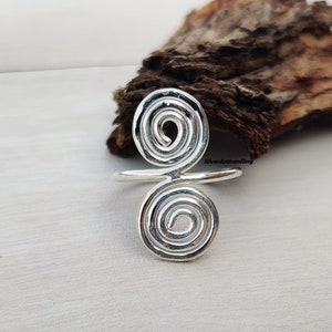 Könnte beinhalten: Ein silberner Ring mit einem Doppelspiralen-Design. Der Ring hat zwei wirbelnde Spiralen, die durch ein Band verbunden sind. Der Ring liegt auf einer hellen Oberfläche mit einem Stück Holz im Hintergrund.