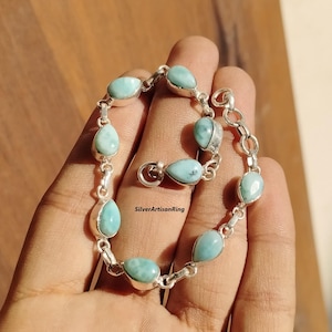 Könnte beinhalten: Ein silbernes Armband mit einem Gliederketten-Design und tropfenförmigen blauen Edelsteinen. Das Armband hat einen Karabinerverschluss.