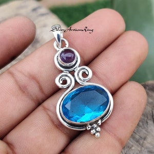 May include: A silver pendant with a blue topaz gemstone and a smaller amethyst gemstone. The pendant has a delicate, ornate design with swirls and a beaded bottom.