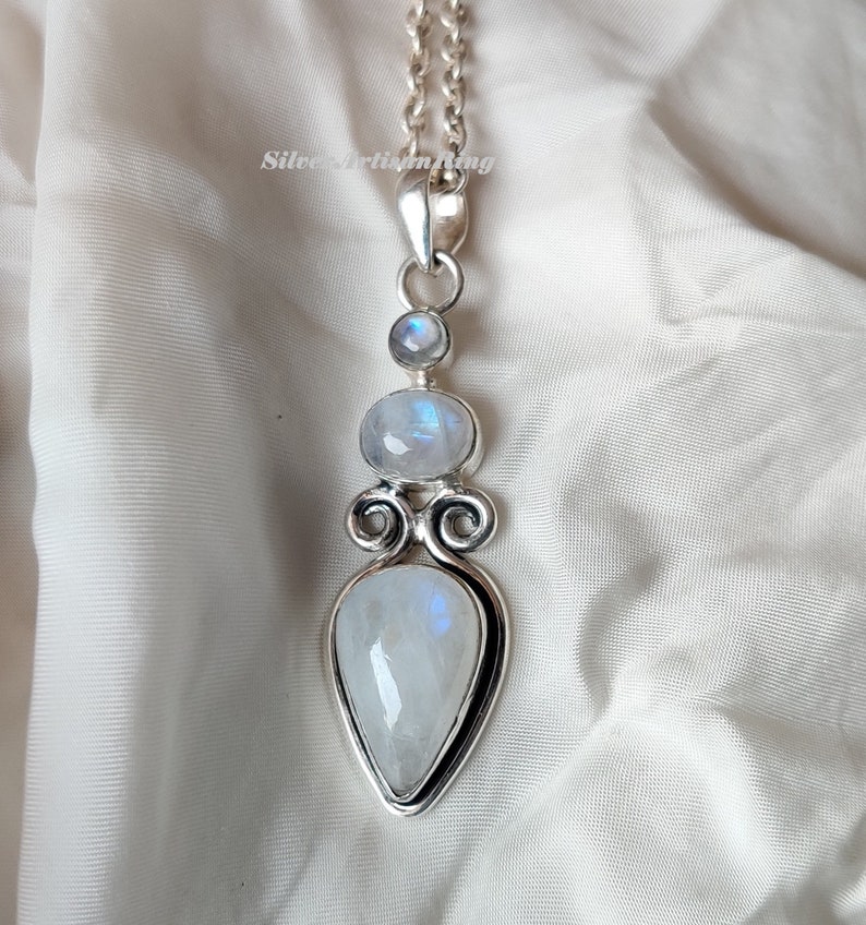 May include: A silver pendant with three moonstone gems. The pendant features a teardrop-shaped moonstone, a round moonstone, and a smaller round moonstone. The pendant is attached to a silver chain.