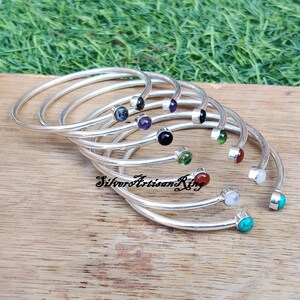 Pode incluir: Um conjunto de pulseiras de prata com pedras preciosas de diferentes cores. As pulseiras est&atilde;o empilhadas umas sobre as outras e as pedras preciosas s&atilde;o vis&iacute;veis. As pulseiras est&atilde;o sobre uma superf&iacute;cie de madeira.