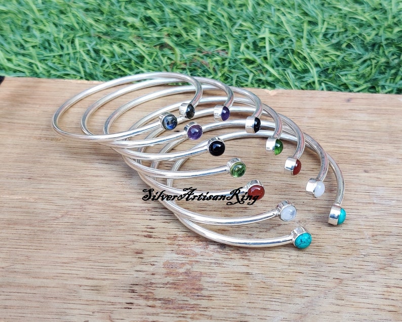 Pode incluir: Um conjunto de pulseiras de prata com pedras preciosas de diferentes cores. As pulseiras est&atilde;o empilhadas umas sobre as outras e as pedras preciosas s&atilde;o vis&iacute;veis. As pulseiras est&atilde;o sobre uma superf&iacute;cie de madeira.