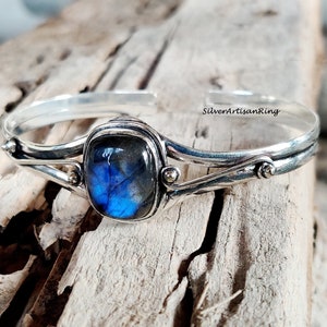 Puede incluir: Un brazalete de plata con una piedra preciosa de labradorita azul engastada en un bisel de plata. El brazalete tiene un dise&ntilde;o delicado y curvo con dos peque&ntilde;os detalles dorados.