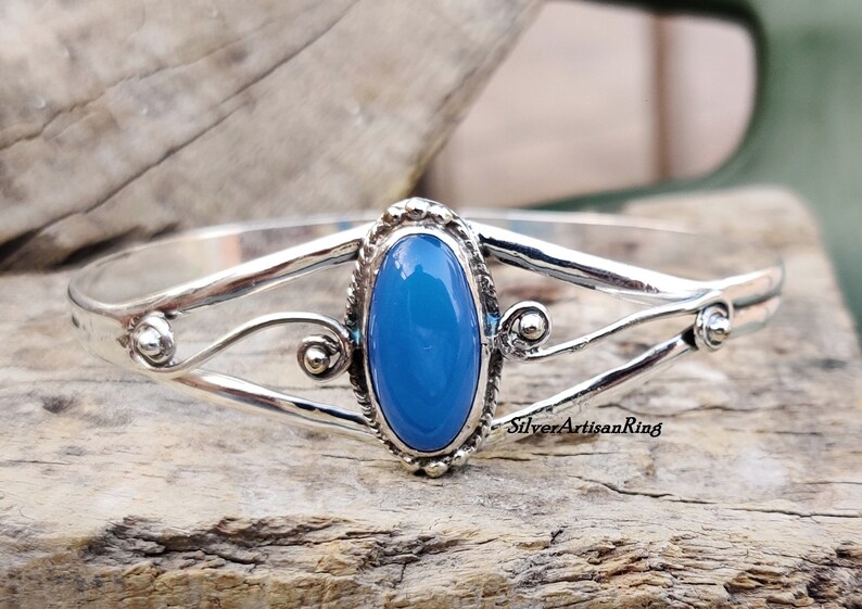 K&ouml;nnte beinhalten: Ein silbernes Armreif-Armband mit einem blauen ovalen Edelstein in der Mitte. Das Armband hat ein zartes, filigranes Design mit Schn&ouml;rkeln und kleinen Perlen.