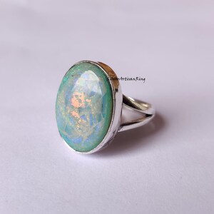 Könnte beinhalten: Ovaler Ring aus Silber mit einem schillernden Edelstein. Der Edelstein zeigt Farbtöne von Hellgrün und Blau mit einem schimmernden Aussehen. Das Band des Rings ist silberfarben und hat ein geteiltes Design. Der Ring ist vor einem weichen, neutralen Hintergrund platziert.