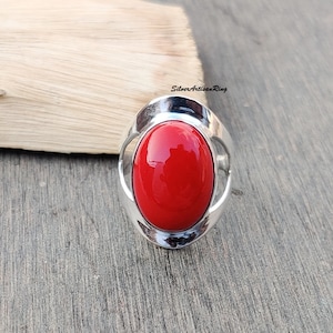 Könnte beinhalten: Ein silberner Ring mit einem großen, ovalen, leuchtend roten Edelstein. Der Ring hat ein schlichtes, modernes Design.