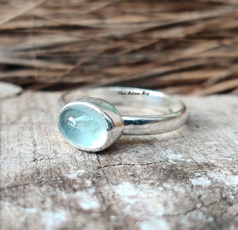 Puede incluir: Un anillo de plata con una piedra ovalada de color azul claro. El anillo est&aacute; colocado sobre una superficie de madera.
