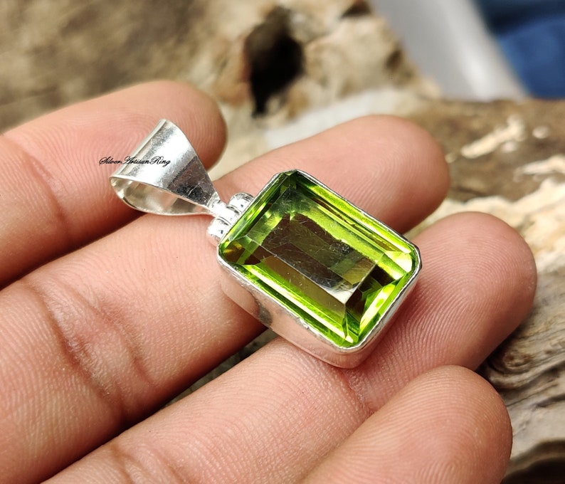 Pode incluir: Um pingente retangular de pedra preciosa verde lim&atilde;o com moldura prateada. O pingente tem um corte retangular e &eacute; colocado em uma moldura de prata polida. O pingente &eacute; segurado em uma m&atilde;o.