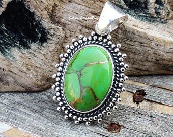 May include: Oval pendant featuring a vibrant green stone with copper-colored veining, set in a silver-toned frame. The frame has a beaded edge and a silver bail for a necklace chain. The pendant is displayed on a wooden surface.