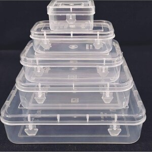 May include: Five clear plastic storage boxes of different sizes are stacked. Each box has a rectangular shape, a hinged lid, and a secure latch. The boxes are arranged in a pyramid, with the largest at the base. The boxes are made of clear plastic.