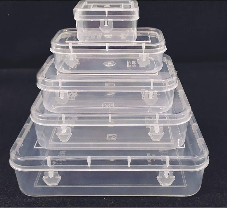 May include: Five clear plastic storage boxes of different sizes are stacked. Each box has a rectangular shape, a hinged lid, and a secure latch. The boxes are arranged in a pyramid, with the largest at the base. The boxes are made of clear plastic.