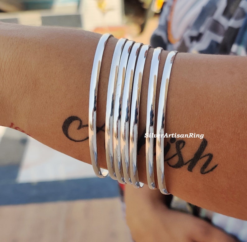 May include: A set of polished silver bangles. The bangles are cylindrical and reflect light. The image displays several bangles worn together. The bangles have a simple, elegant design, suitable for everyday wear.