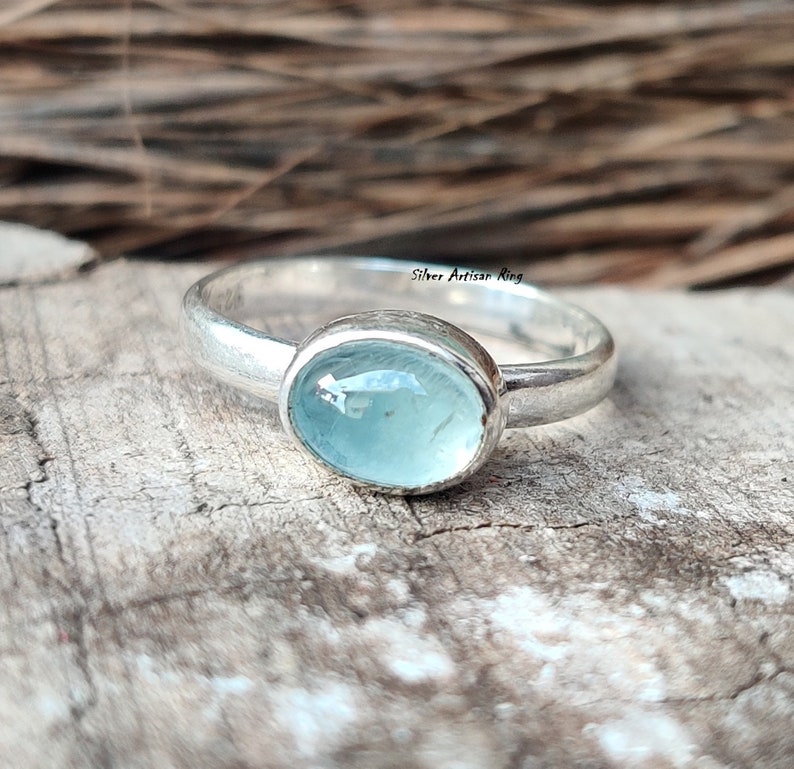 Puede incluir: Un anillo de plata con una piedra ovalada azul claro. El anillo est&aacute; sobre una superficie de madera. "Silver Artisan Ring" est&aacute; escrito en el anillo.