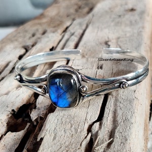 Puede incluir: Un brazalete de plata con una piedra preciosa de labradorita azul engastada en un engaste de plata. El brazalete tiene un dise&ntilde;o delicado y retorcido y presenta peque&ntilde;as cuentas de plata.