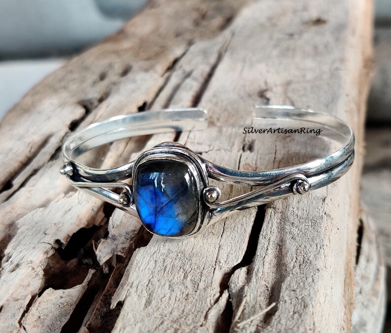Puede incluir: Un brazalete de plata con una piedra preciosa de labradorita azul engastada en un engaste de plata. El brazalete tiene un dise&ntilde;o delicado y retorcido y presenta peque&ntilde;as cuentas de plata.