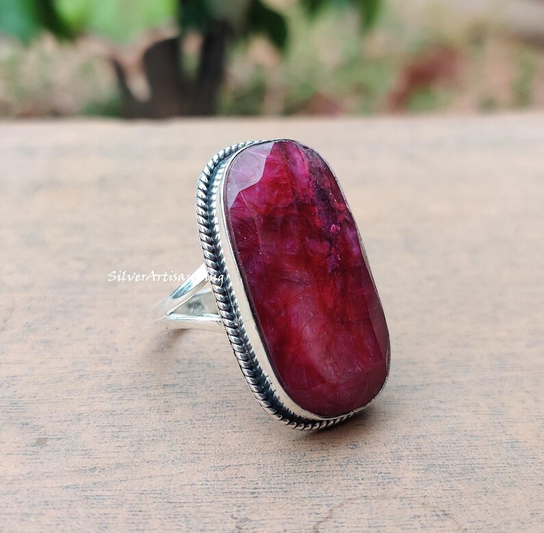 May include: A silver ring with a large, oval ruby gemstone. The ruby is a deep red hue and is set in a bezel setting with a rope-like border. The ring has a polished finish and is a statement piece.