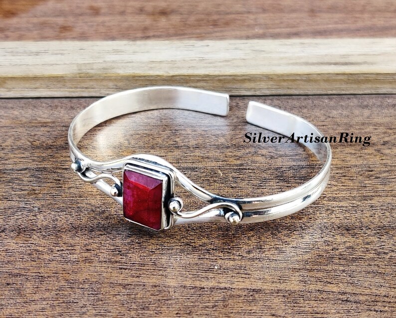 May include: A silver bangle bracelet featuring a red gemstone set in a square bezel. The bracelet has a delicate, wavy design and is open on one side.