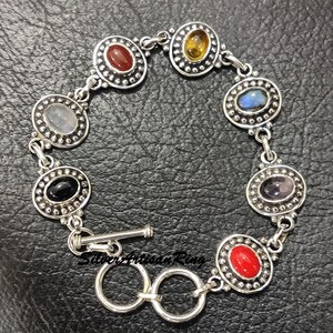 May include: A silver bracelet with seven oval-shaped gemstones set in silver bezels. The gemstones are red, orange, yellow, green, blue, purple, and black. The bracelet has a toggle clasp.