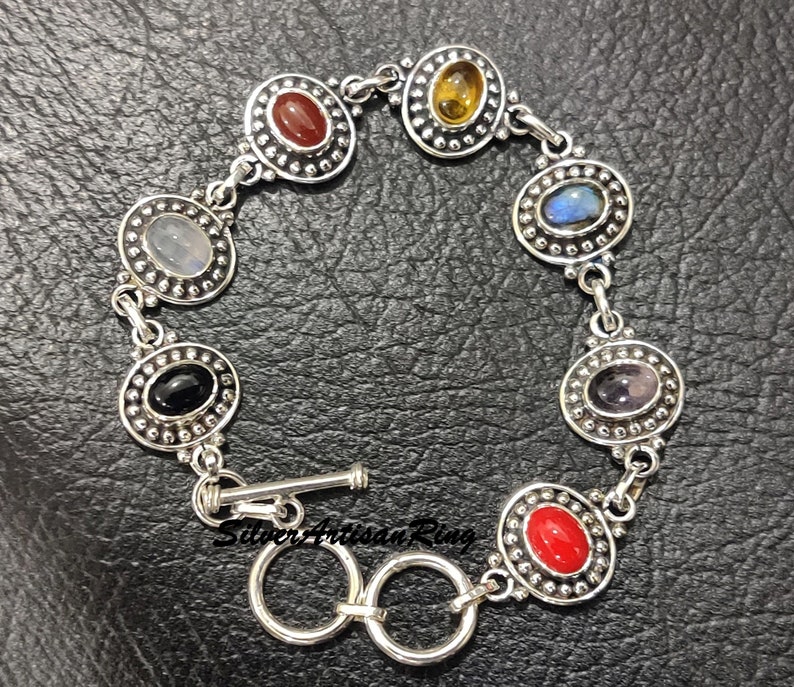 May include: A silver bracelet with seven oval-shaped gemstones set in silver bezels. The gemstones are red, orange, yellow, green, blue, purple, and black. The bracelet has a toggle clasp.