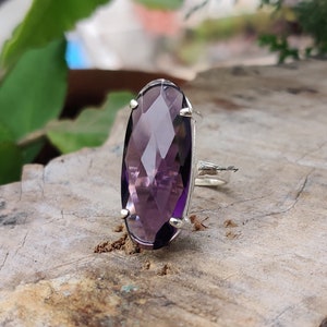 May include: A silver ring with a large, faceted, oval-shaped amethyst gemstone. The ring is set on a wooden surface.