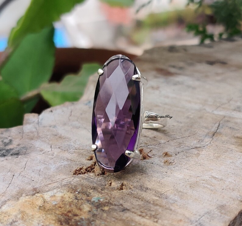 May include: A silver ring with a large, faceted, oval-shaped amethyst gemstone. The ring is set on a wooden surface.