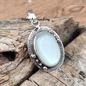 Op de afbeelding: Een zilveren hanger met een witte ovale edelsteen, gezet in een getextureerd zilveren frame. Het frame is versierd met kleine zilveren kralen rond de rand.