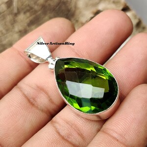 May include: A pear-shaped green gemstone pendant with a silver setting. The pendant is held in a hand.