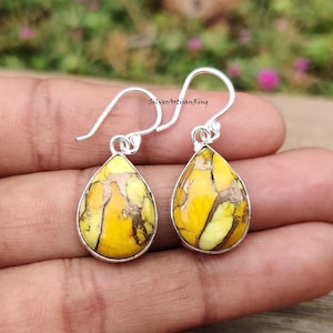 May include: A pair of silver drop earrings with teardrop-shaped yellow and brown stone pendants. The earrings have a hook closure and are set in a silver bezel. The stone has a marbled appearance.