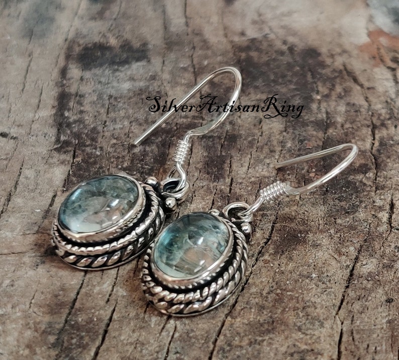 May include: Silver earrings with a blue gemstone set in a silver bezel with a rope-like design. The earrings have a hook closure.