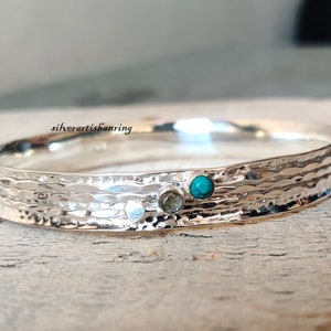May include: Silver bangle bracelet with a hammered texture and two small gemstones. The larger gemstone is turquoise blue and the smaller gemstone is a light brown color.